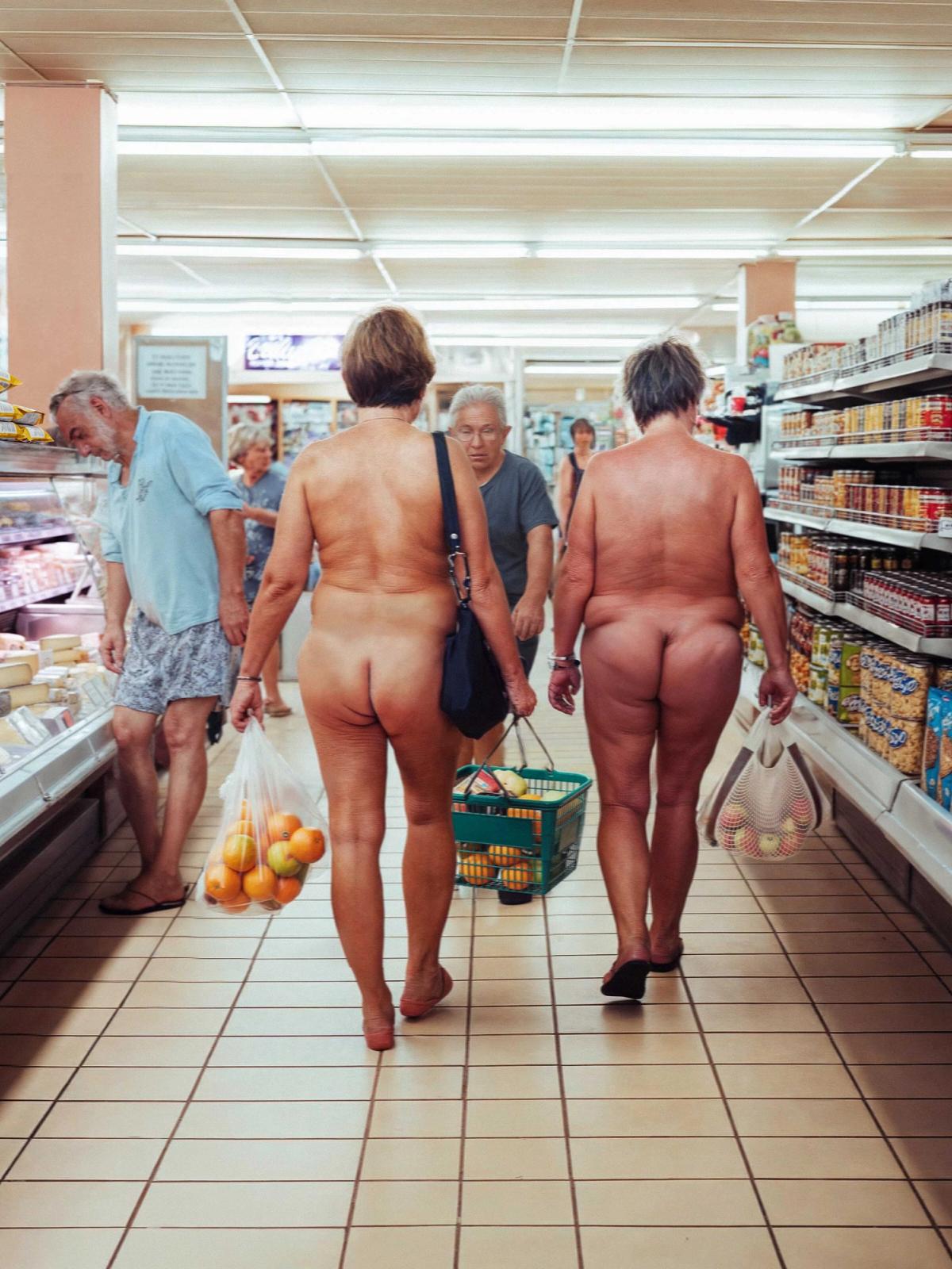 The image shows the interior of a grocery store from a low-angle perspective behind two naked women walking down the aisle. One woman carries a plastic bag of oranges and a black purse, while the other carries a mesh bag of groceries. Other shoppers, some clothed and some naked, are visible further down the aisle near shelves stocked with snacks and produce.