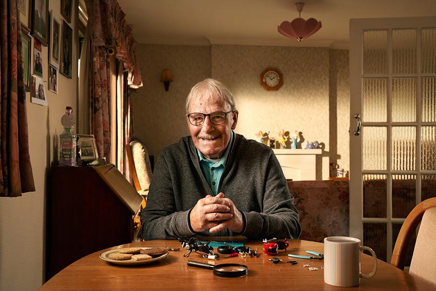 An older man with white hair and a grey cardigan sits at a table with a plate of biscuits and a cup of coffee, surrounded by various small objects and tools.