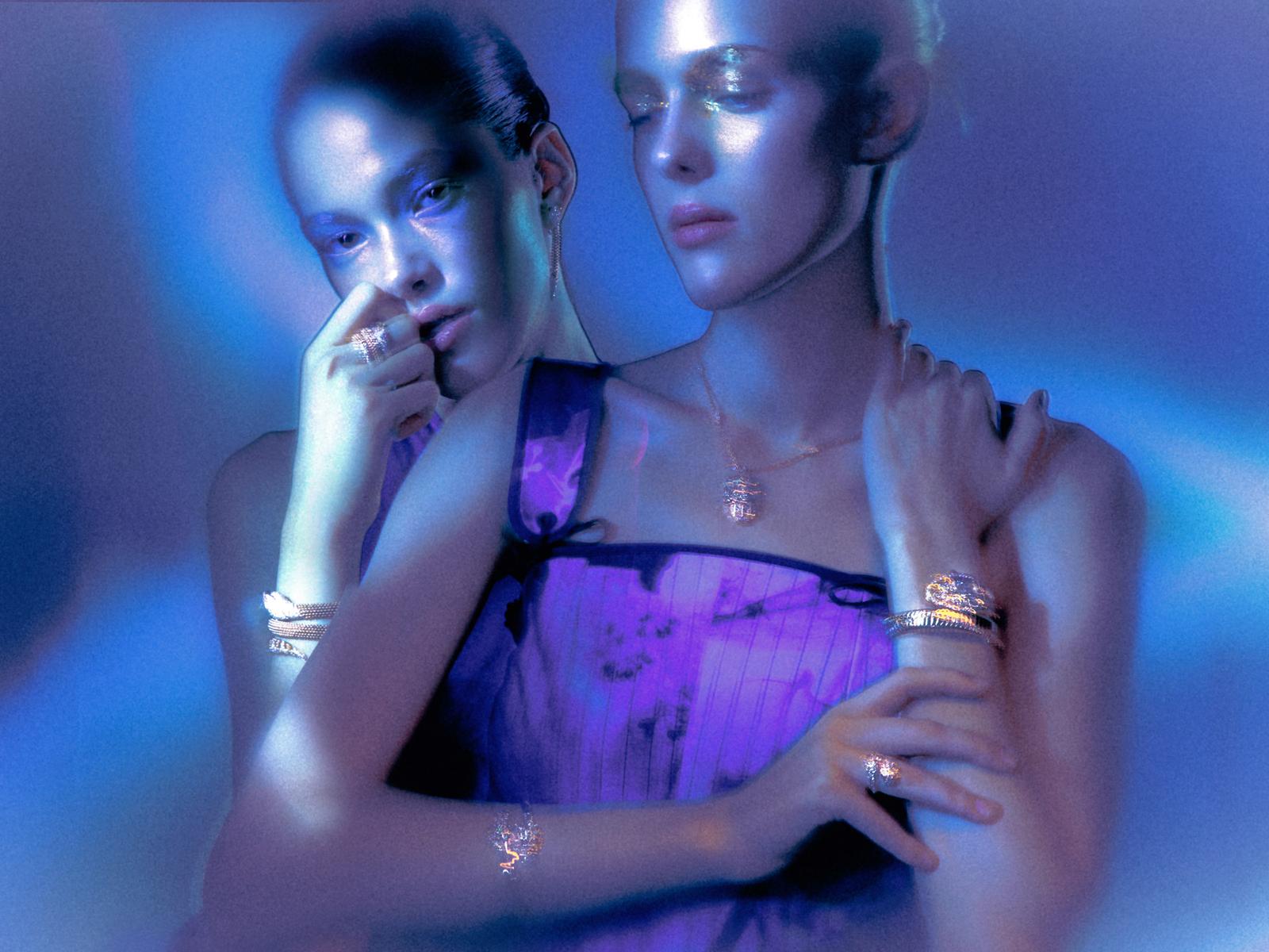 Two women in matching purple dresses, one with her hand on the other's shoulder, posing in a dimly lit, blue-lit room.