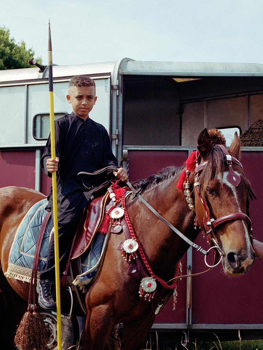 A young boy in a black shirt and pants, holding a spear, riding a brown horse in front of a red and white horse trailer.