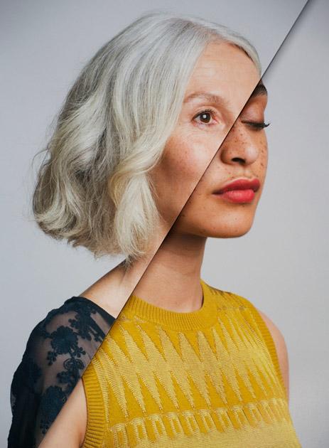 A woman with short, platinum blonde hair and a yellow sweater with a black lace sleeve, looking directly at the camera with a neutral expression.