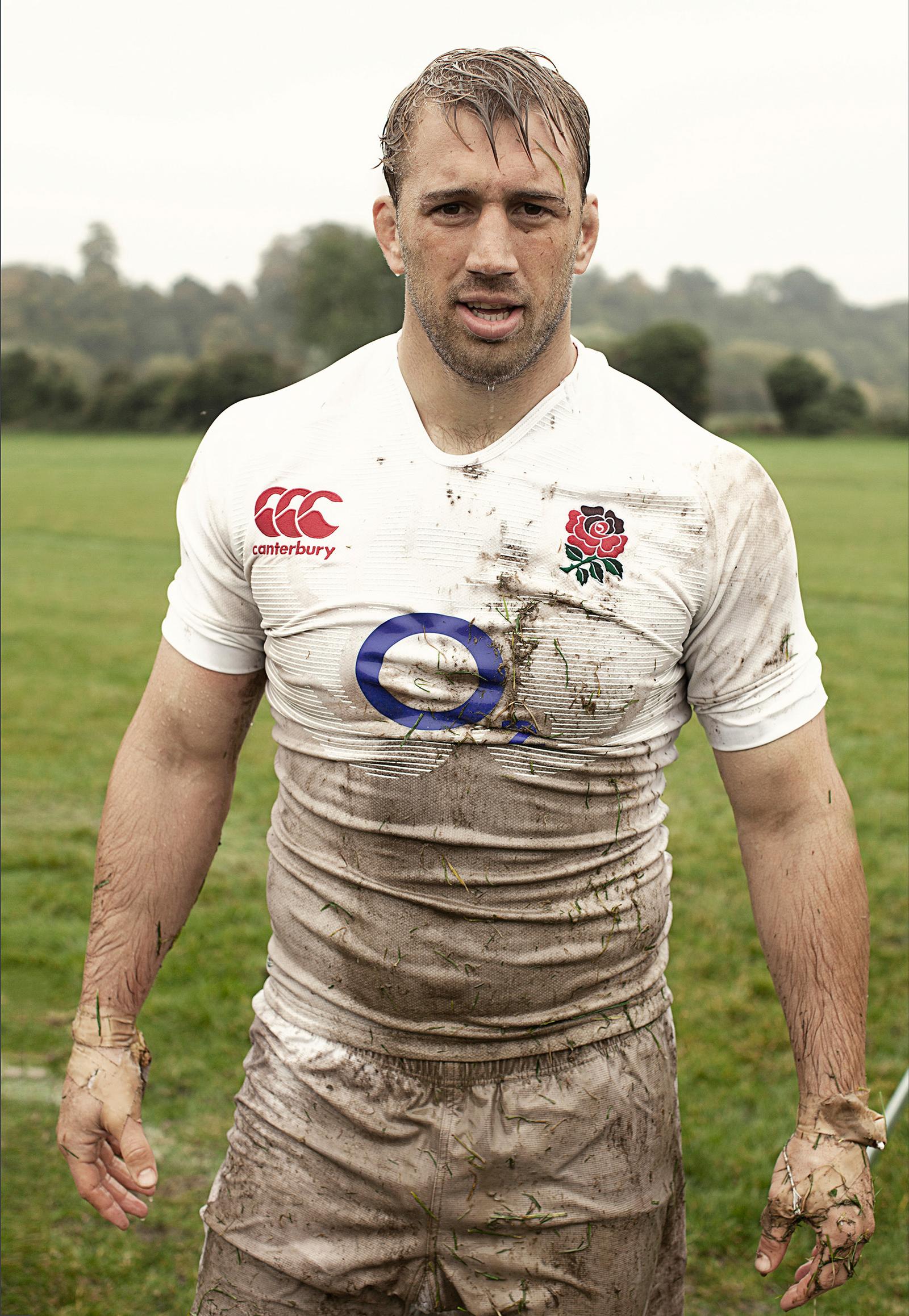 A man covered in mud and grass, wearing a white rugby jersey with a red rose emblem on the left chest, and white rugby shorts, standing on a grassy field.