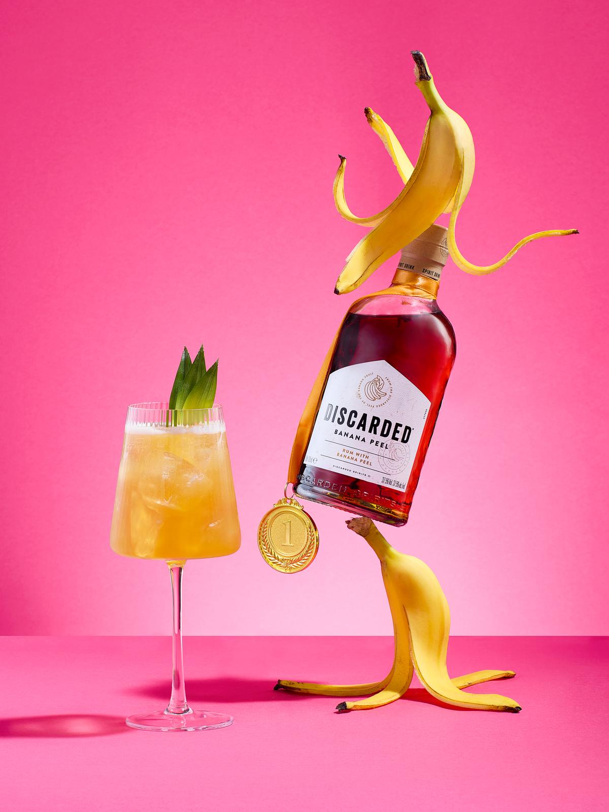 A cocktail glass containing a yellow, orange, and white drink, garnished with a pineapple leaf, next to a bottle of rum with a banana peel label, on a pink background.