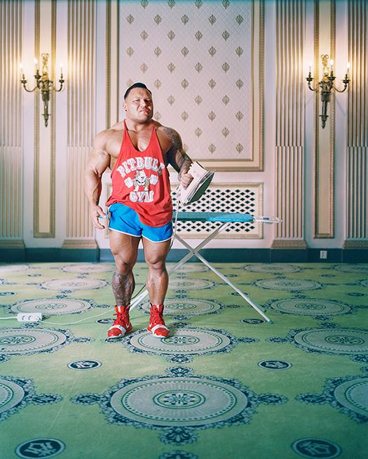 A man in a red tank top and blue shorts is standing in a room with a large, ornate wall behind him, holding an ironing board with an iron in his hand.