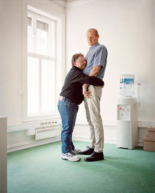 A man and a woman embracing in an office setting, with the man wearing a blue shirt and khaki pants, and the woman wearing a black shirt and blue jeans.