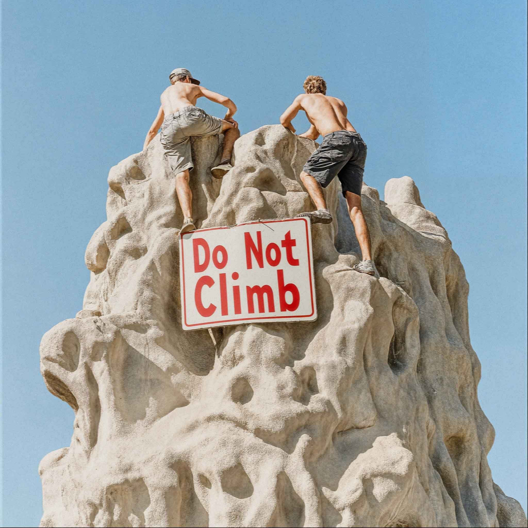 Two men in shorts and no shirts climbing a large rock formation with a 'Do Not Climb' sign in the foreground.
