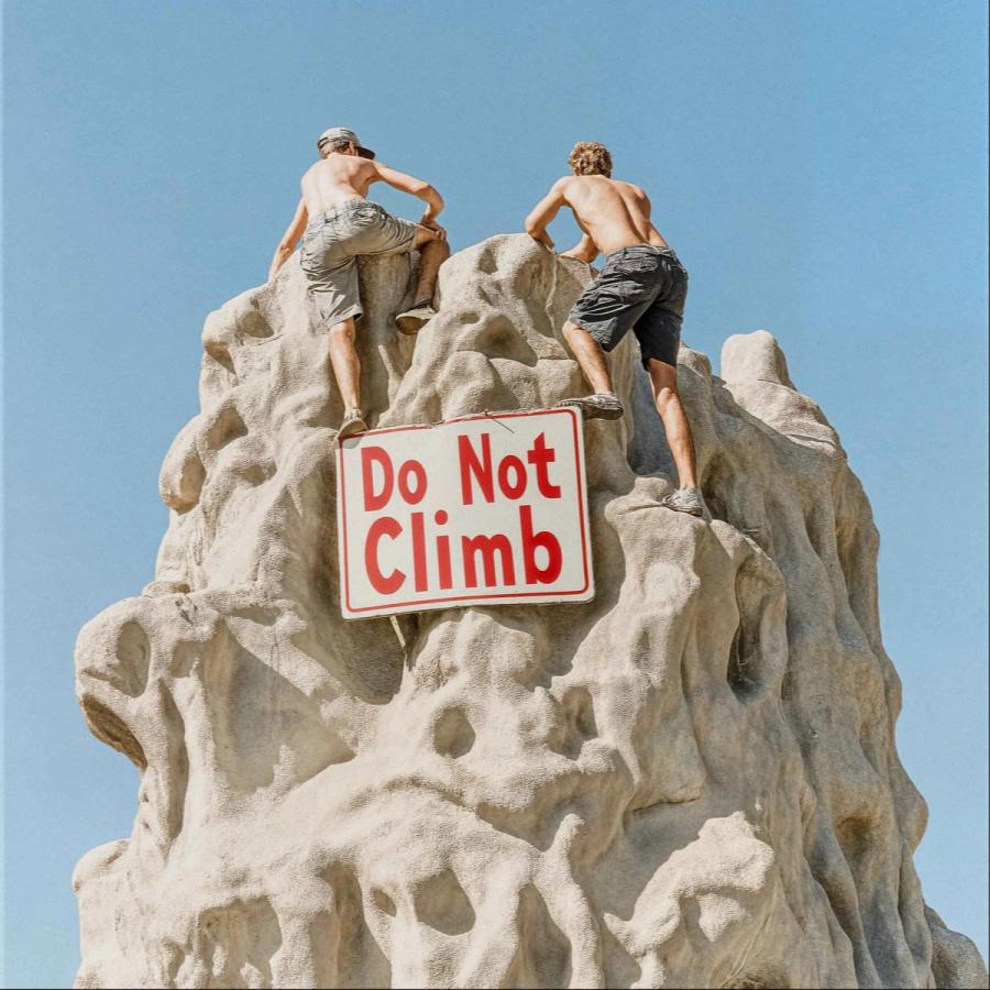 Two men in shorts and no shirts climbing a large rock formation with a 'Do Not Climb' sign in the foreground.