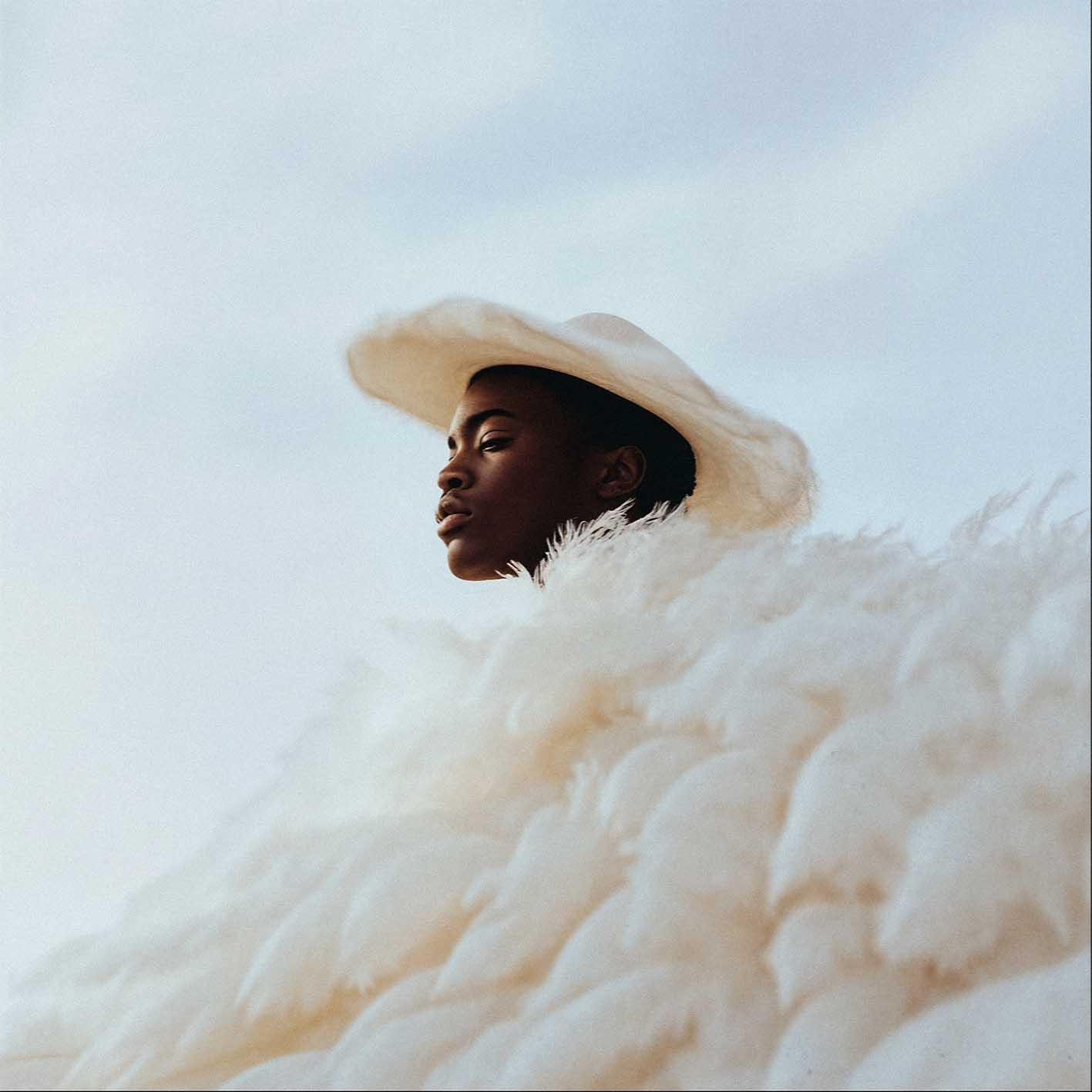A woman wearing a white cowboy hat and a white fur coat, standing in a desert landscape.