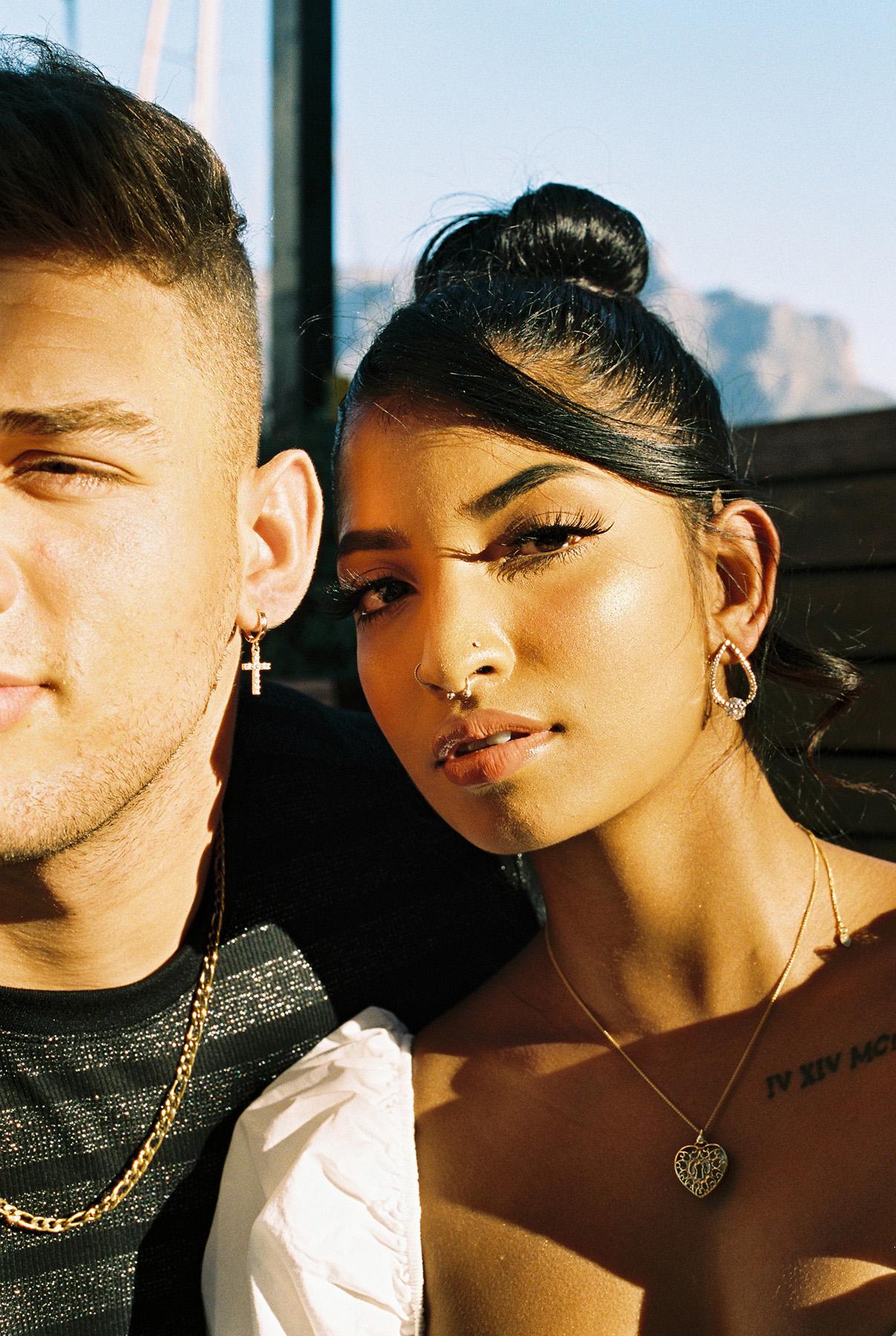A man and a woman with dark skin and hair, both wearing gold jewelry and looking directly at the camera.