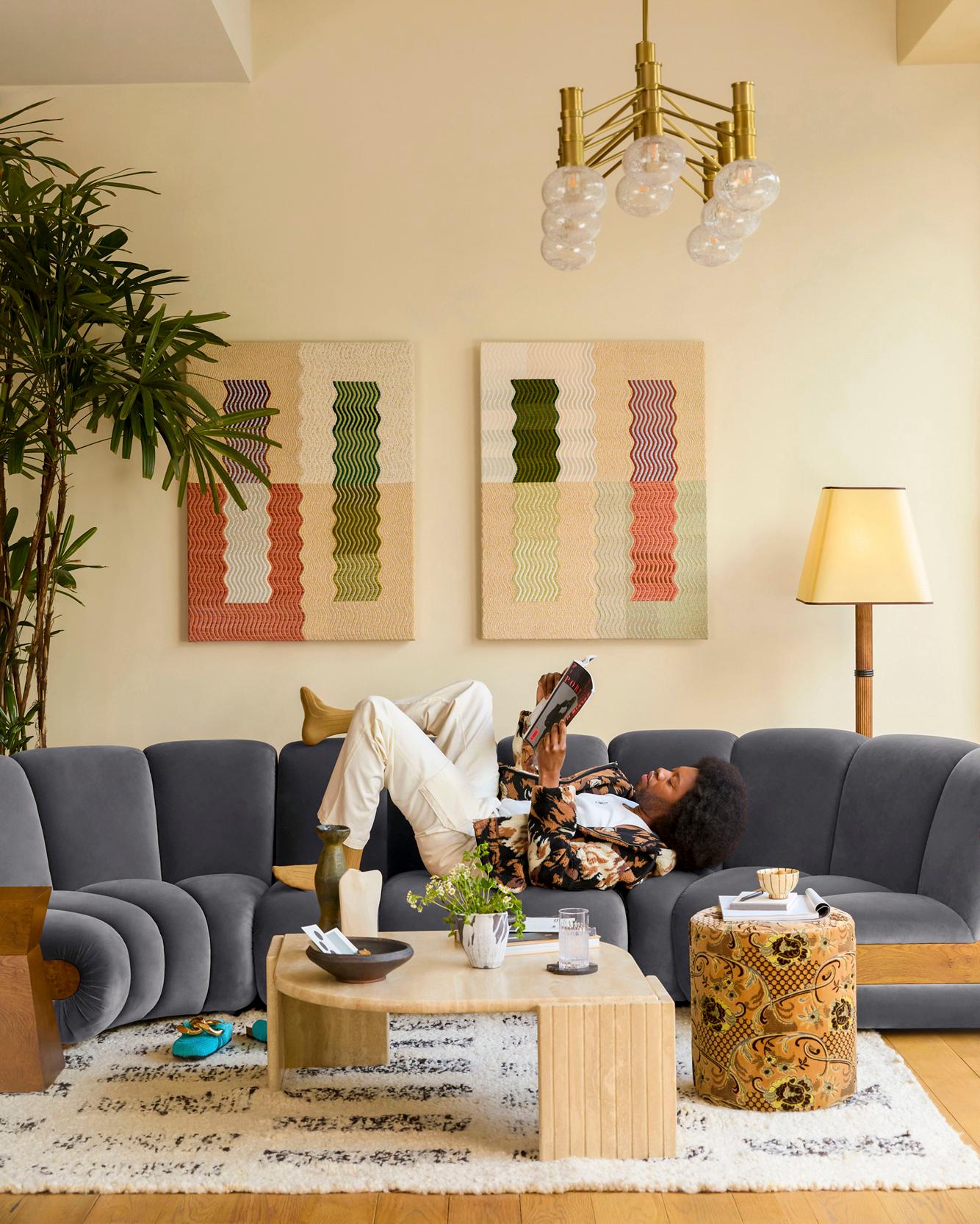 A person is lying on a gray couch, reading a book, with a coffee table in front of them, and a plant in the corner.