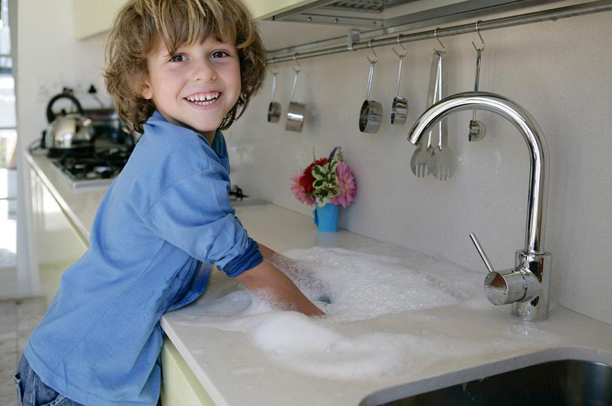 A young boy with messy brown hair and a blue shirt is washing his hands in a kitchen sink, surrounded by soap suds and water.