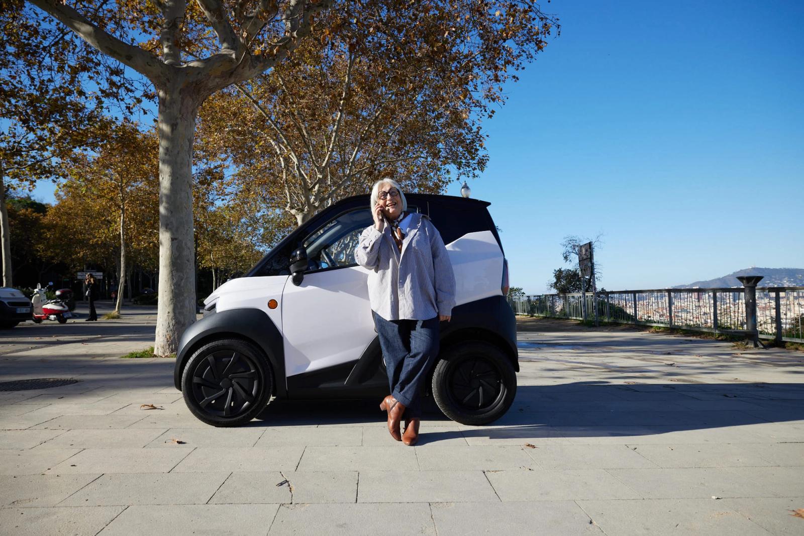 A woman stands in front of a white electric car, smiling and holding her phone to her ear. She has short white hair and is dressed in a light gray sweater and jeans, standing on.