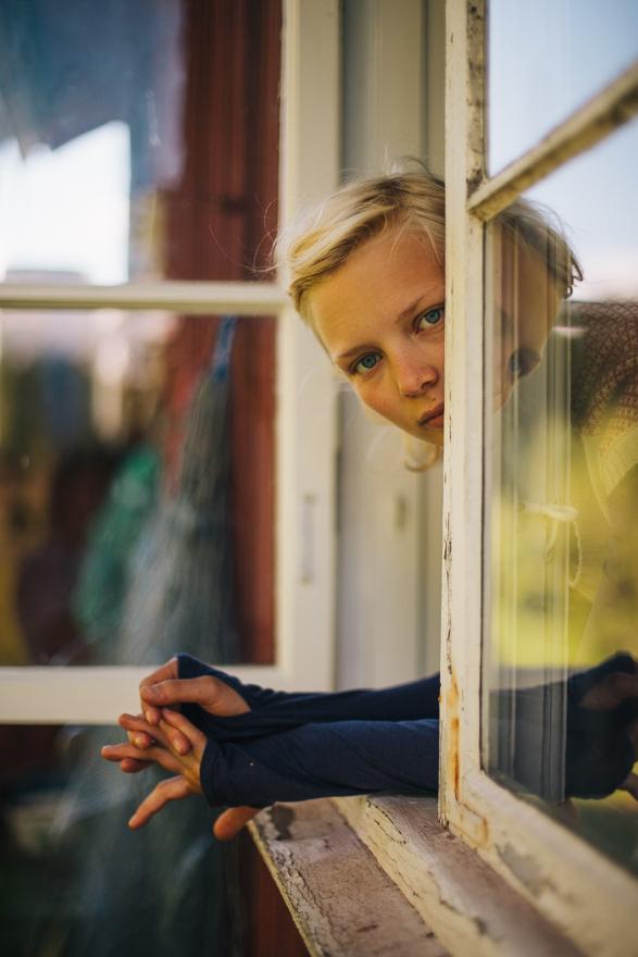 A young girl with blonde hair and blue eyes peering out from behind a window frame, wearing a brown sweater.