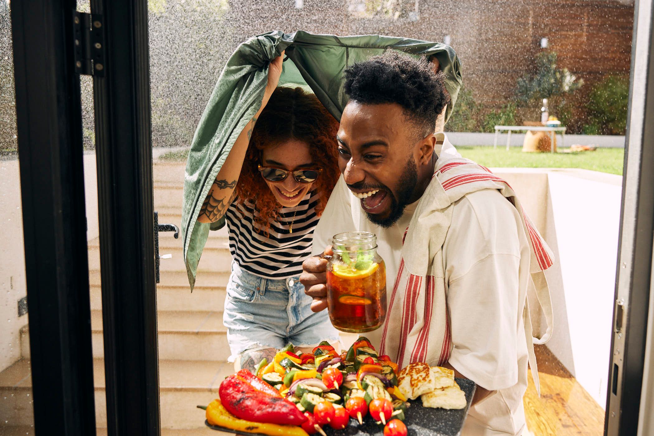 A man and woman are laughing and covering their heads with a green blanket as they exit a house with a tray of food. The woman has red hair and a striped shirt, while the man has.