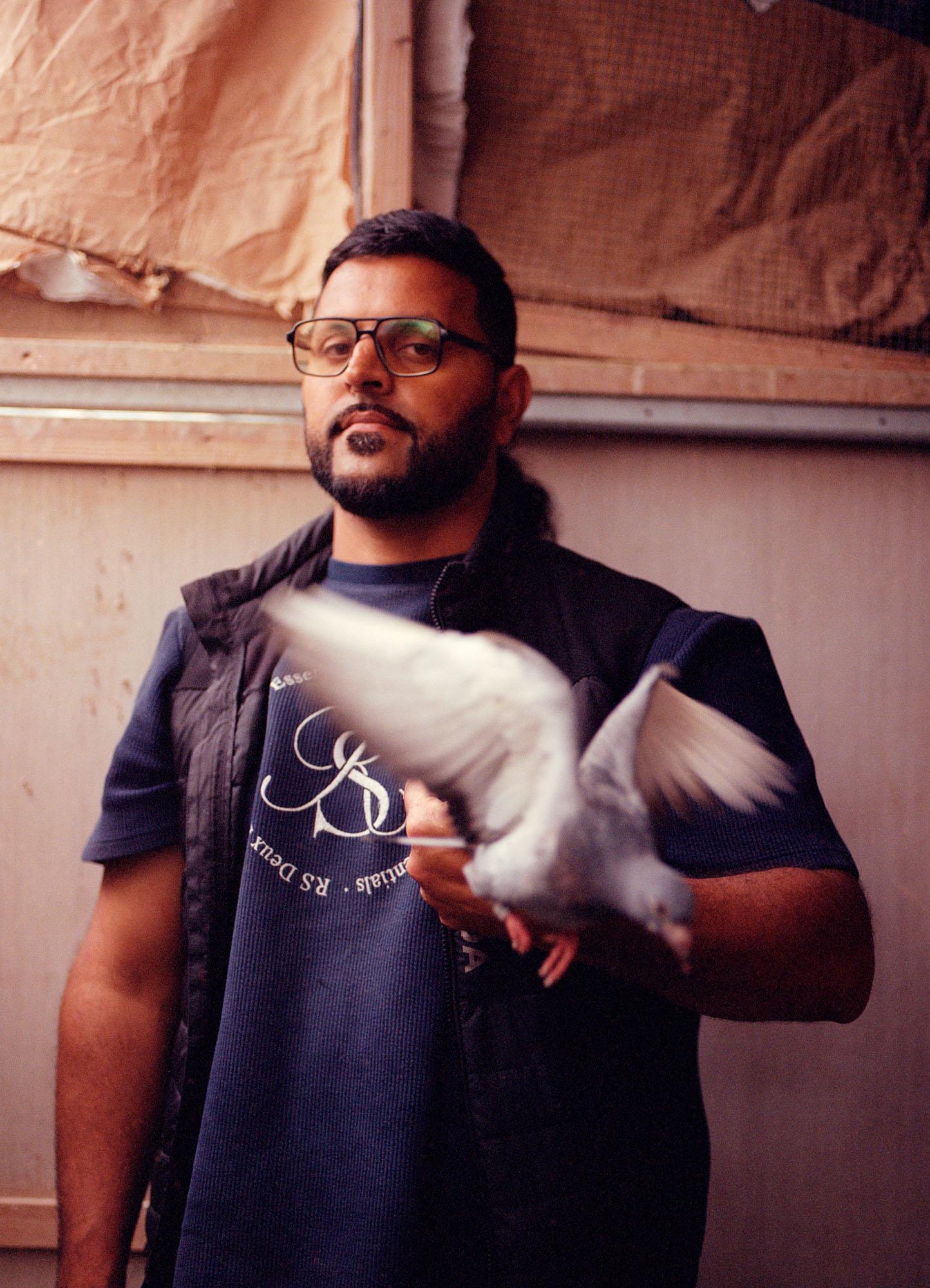 A man with a beard and glasses holding a white pigeon in a studio setting, wearing a blue t-shirt and a black vest.