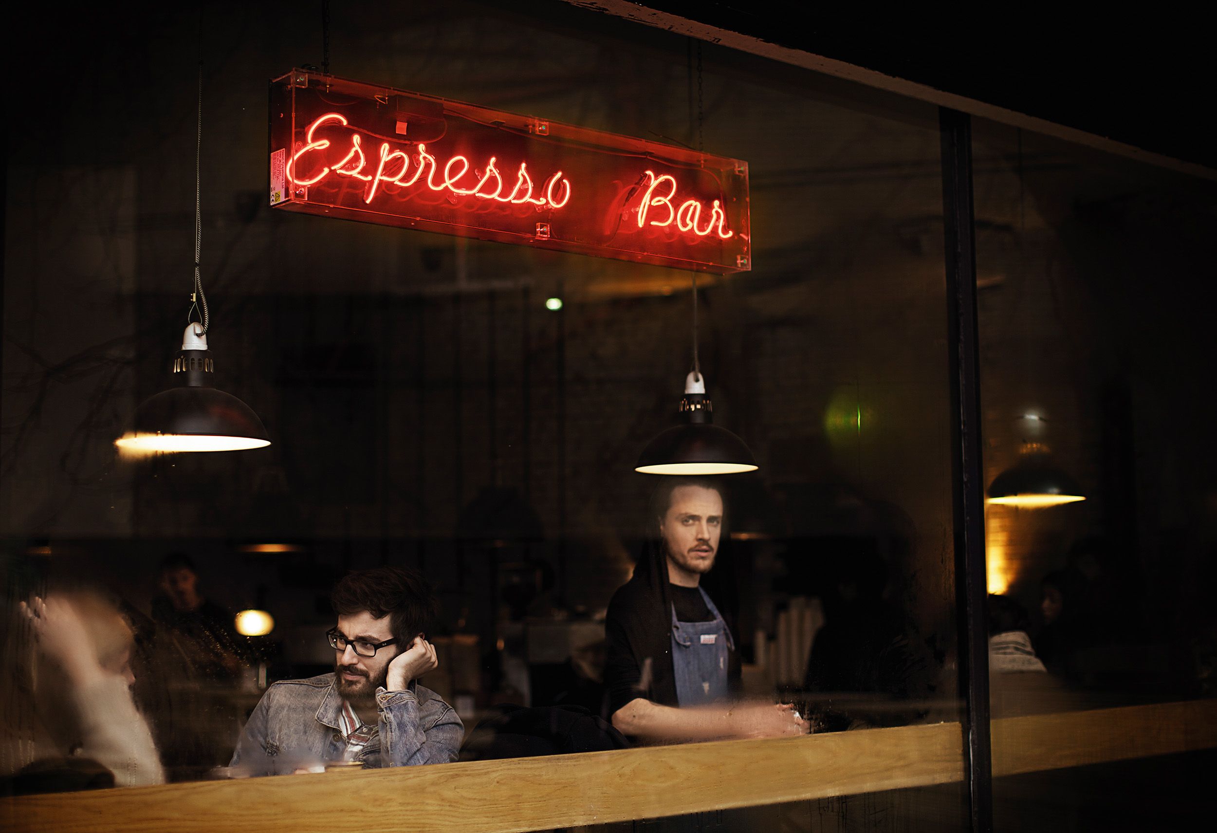 A man in a denim jacket sits at a table while looking out a coffee shop window at another man behind the counter.