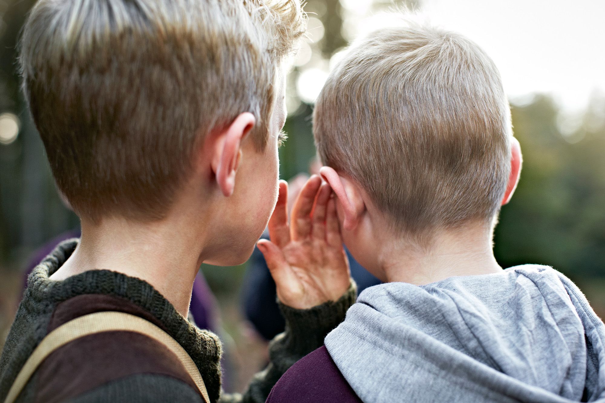 Two people, likely a mother and child, are sharing a secret in a quiet outdoor setting, with the mother placing her hand over the child's ear.