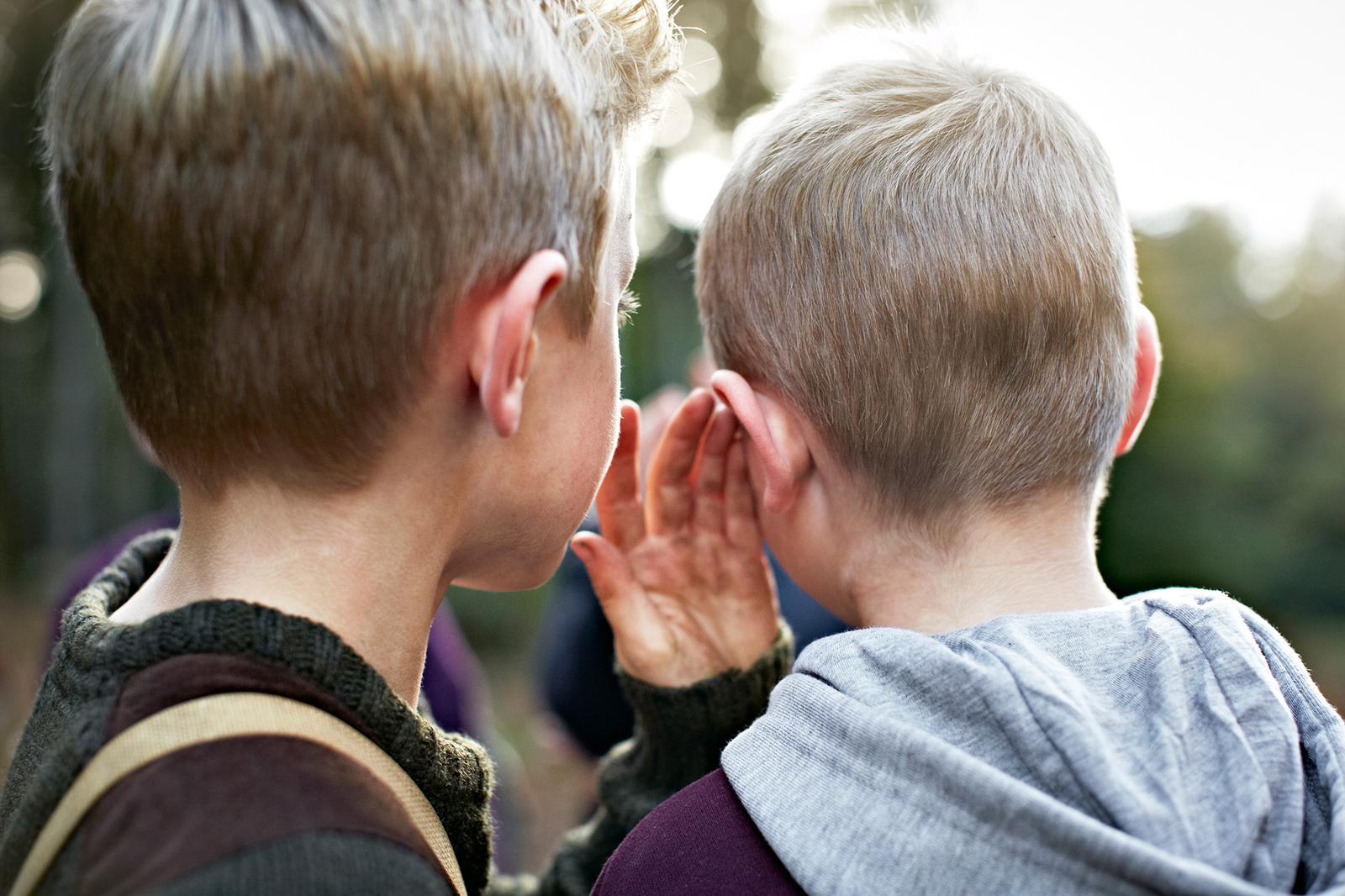 Two people, likely a mother and child, are sharing a secret in a quiet outdoor setting, with the mother placing her hand over the child's ear.