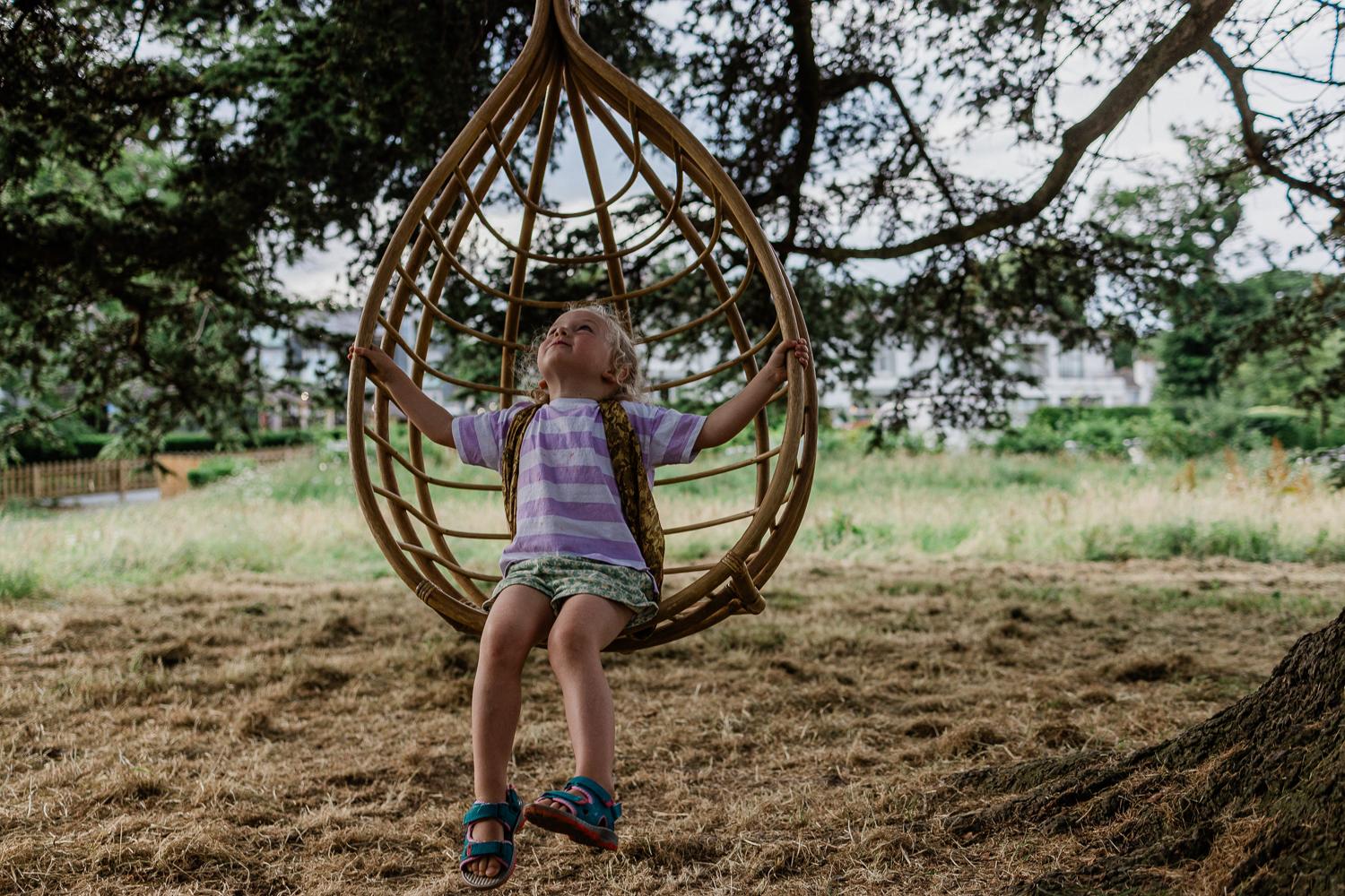 A young girl is sitting in a large, wooden, teardrop-shaped swing, suspended from a tree branch, in a lush, green, outdoor setting.