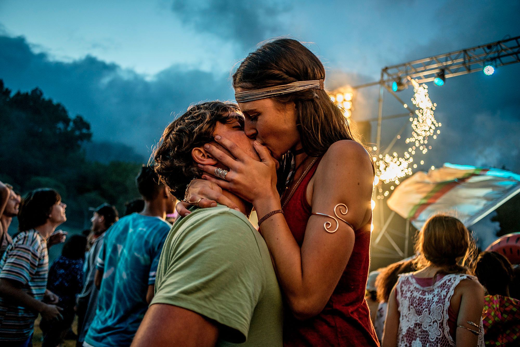 A young woman in a red dress kissing a man in a green t-shirt at a music festival or outdoor event, with a crowd of people in the background.