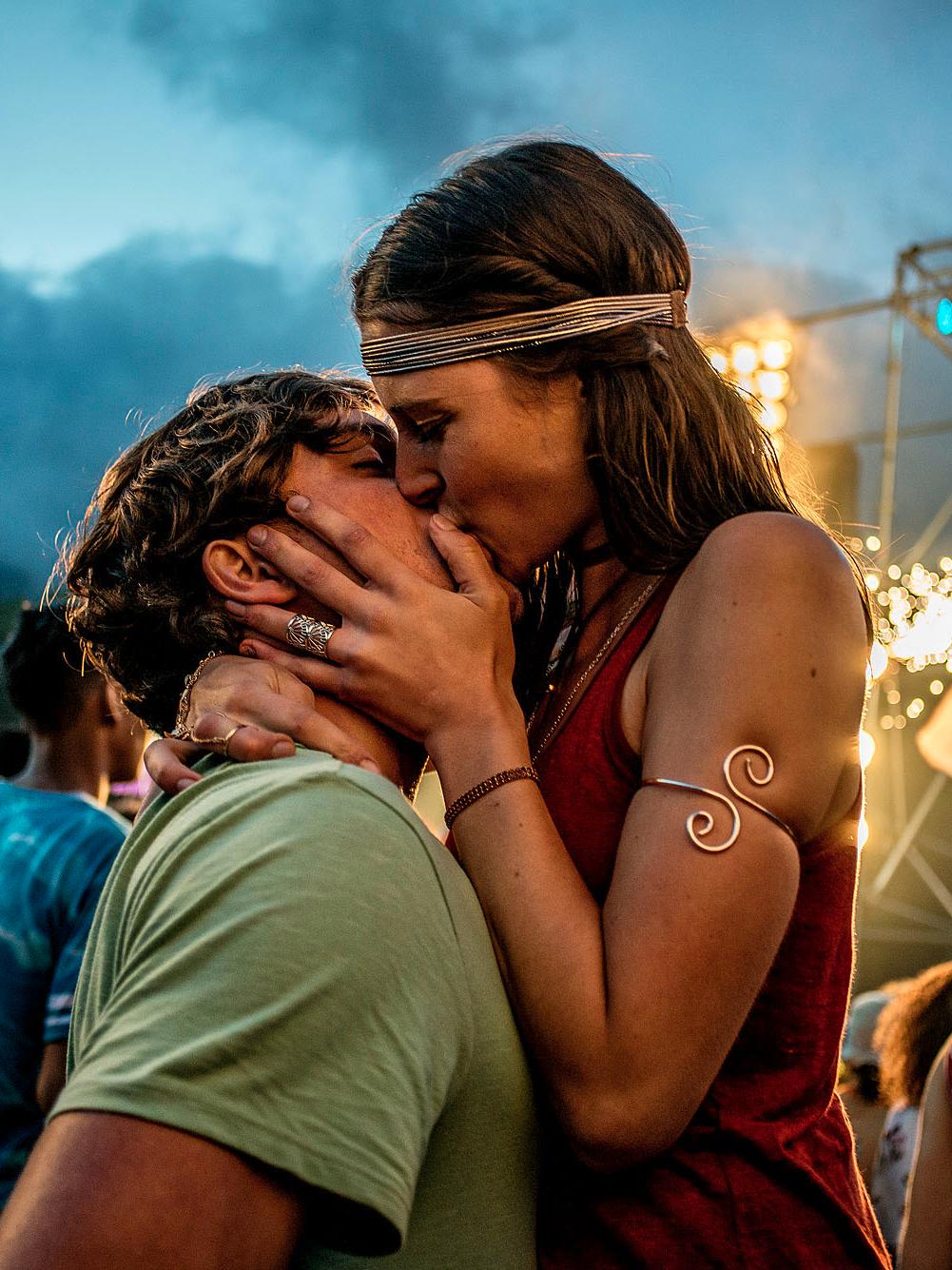 A young woman in a red dress kissing a man in a green t-shirt at a music festival or outdoor event, with a crowd of people in the background.