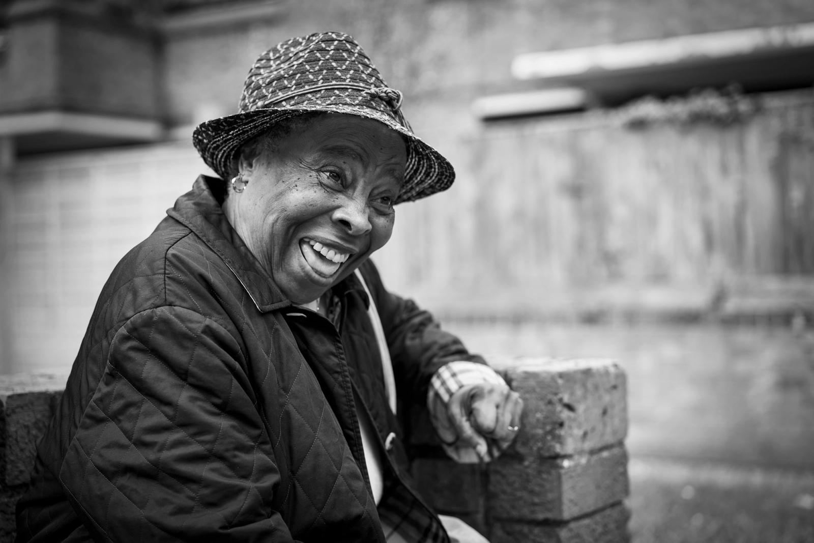 An older woman with a wide smile, wearing a patterned bucket hat, a dark quilted jacket, and a plaid shirt, sitting on a brick wall.