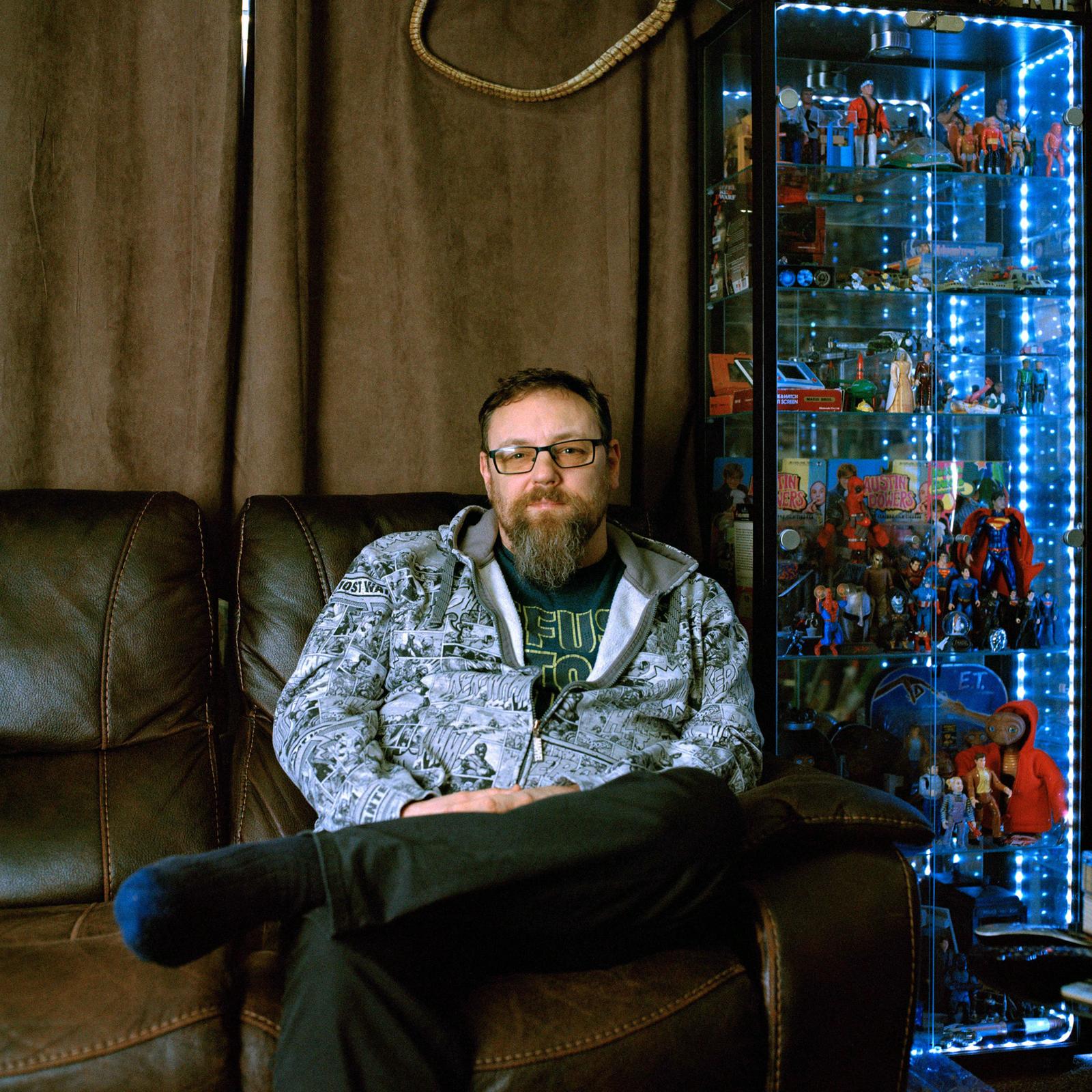 A man sits in a chair with a beard and glasses, surrounded by action figures and toys in a glass cabinet.