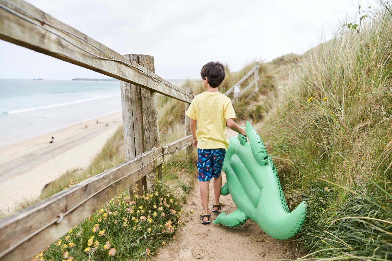 A young boy, dressed in a yellow t-shirt and blue shorts with a pattern of red and white shapes, walks along a sandy path with a green inflatable crocodile. The boy is holding.