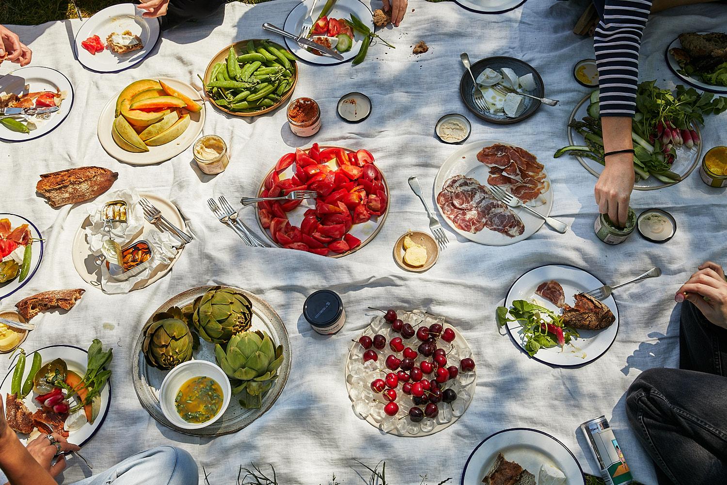 A group of people are having a picnic on a blanket in a park, with a variety of food and drinks spread out in front of them.