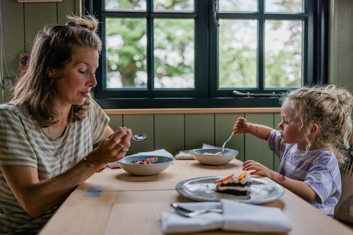 A woman and a young girl eating dessert at a table in a restaurant, with a window in the background showing a tree outside.