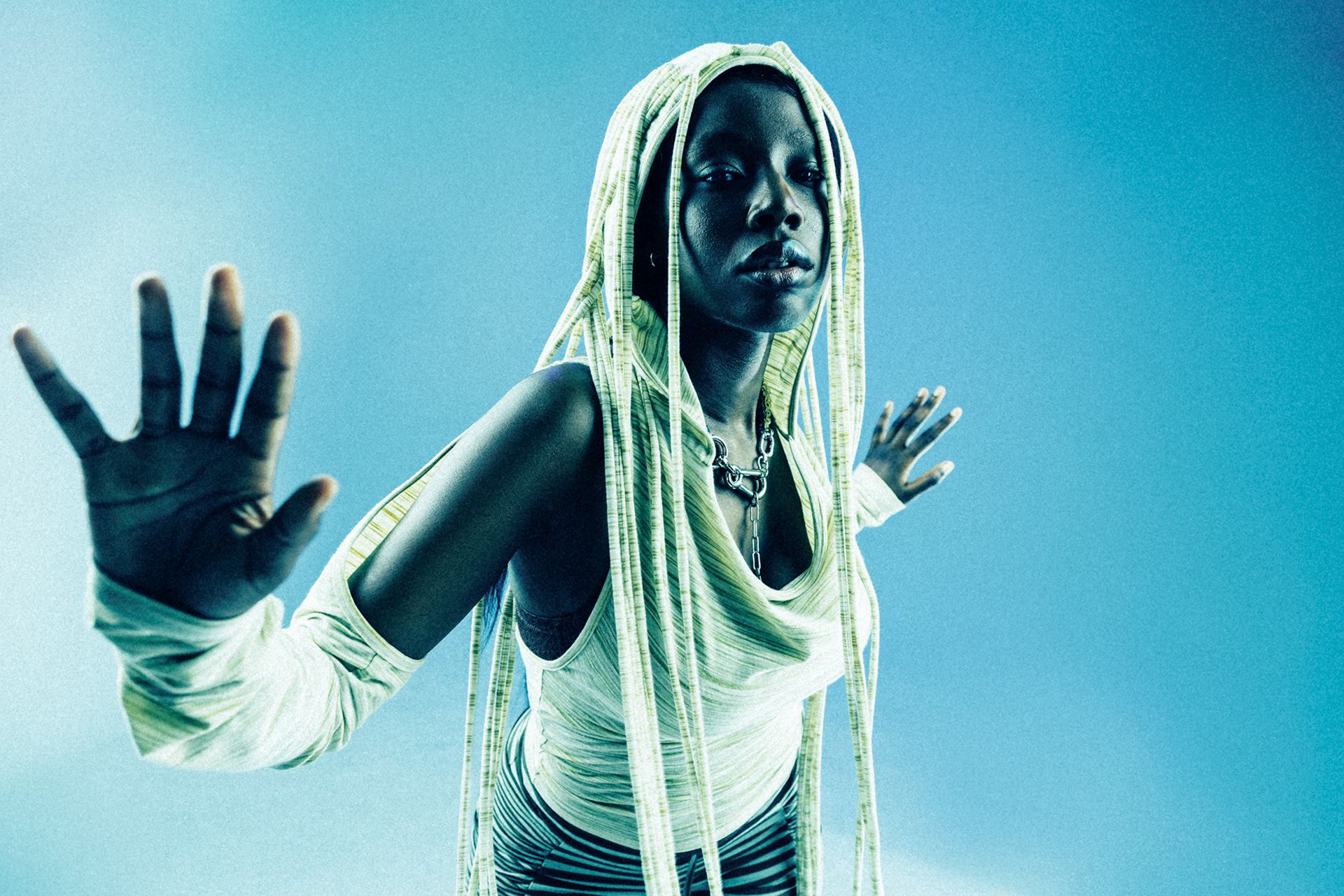 A woman with long, braided hair and a white, sleeveless top with a hood, standing in a dark, urban environment with a cityscape in the background.