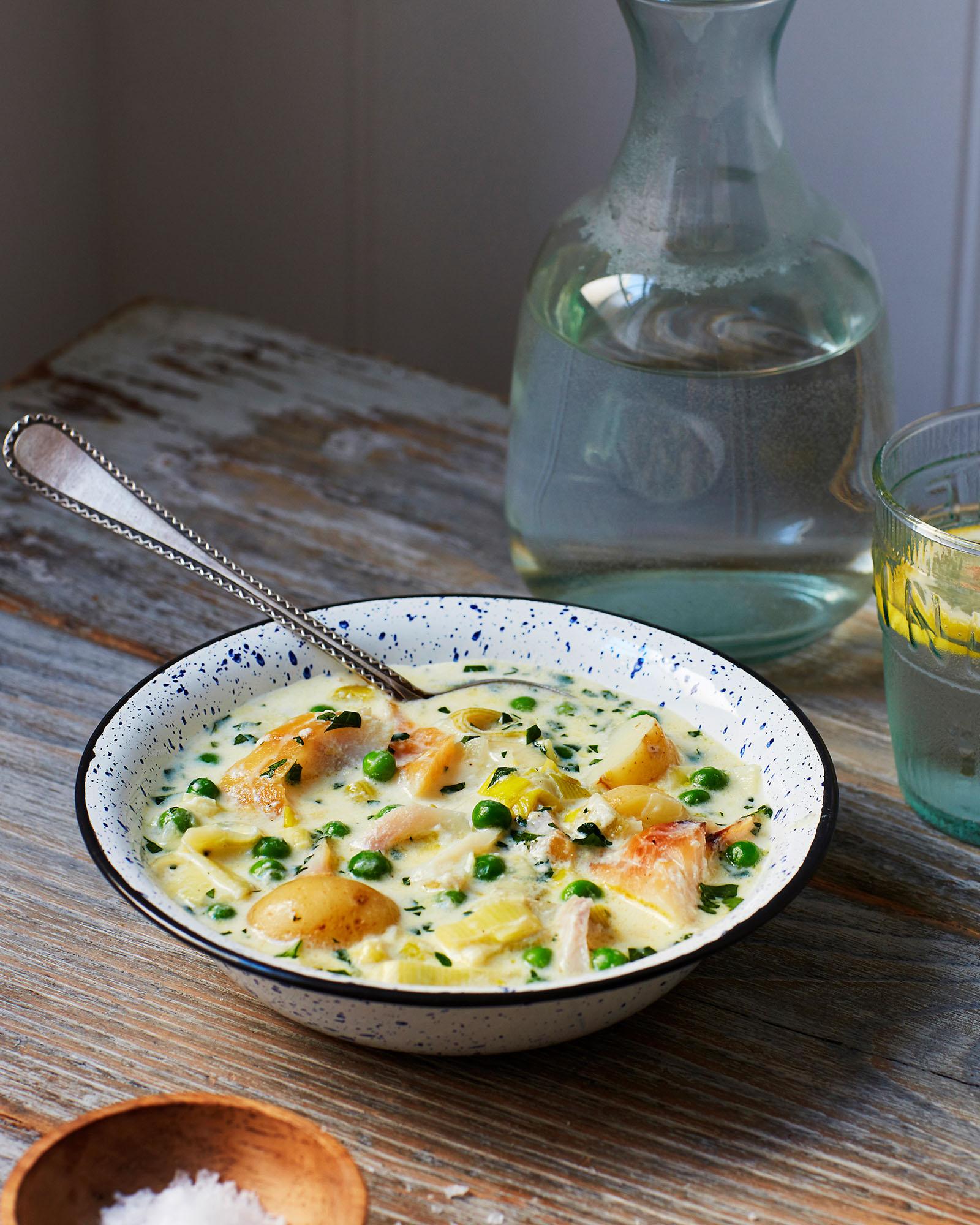 A bowl of creamy fish and vegetable stew, with a spoon in it, on a wooden table.