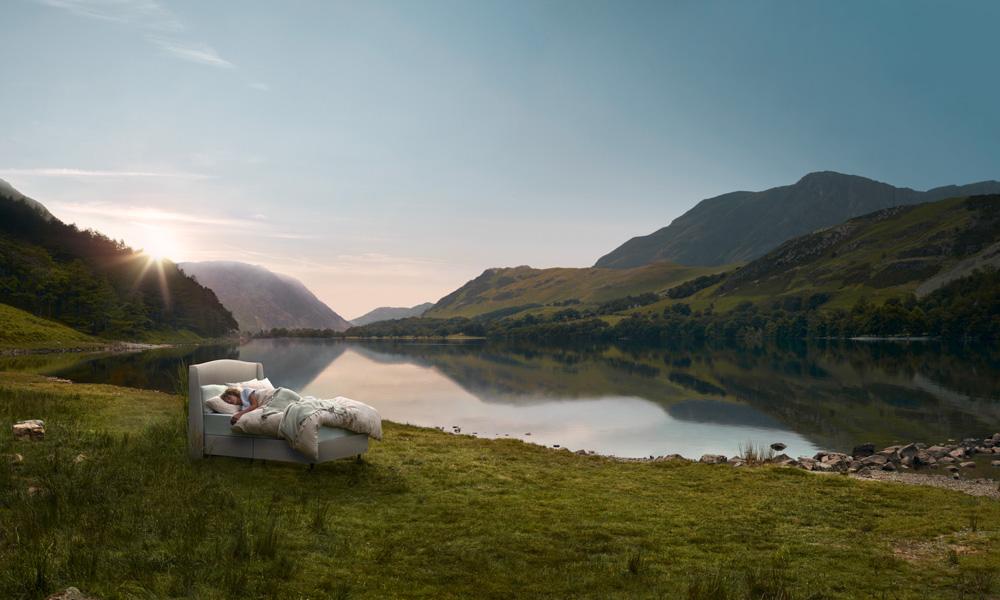 Two people are sleeping in a bed outdoors, surrounded by a serene lake and mountains at sunset.