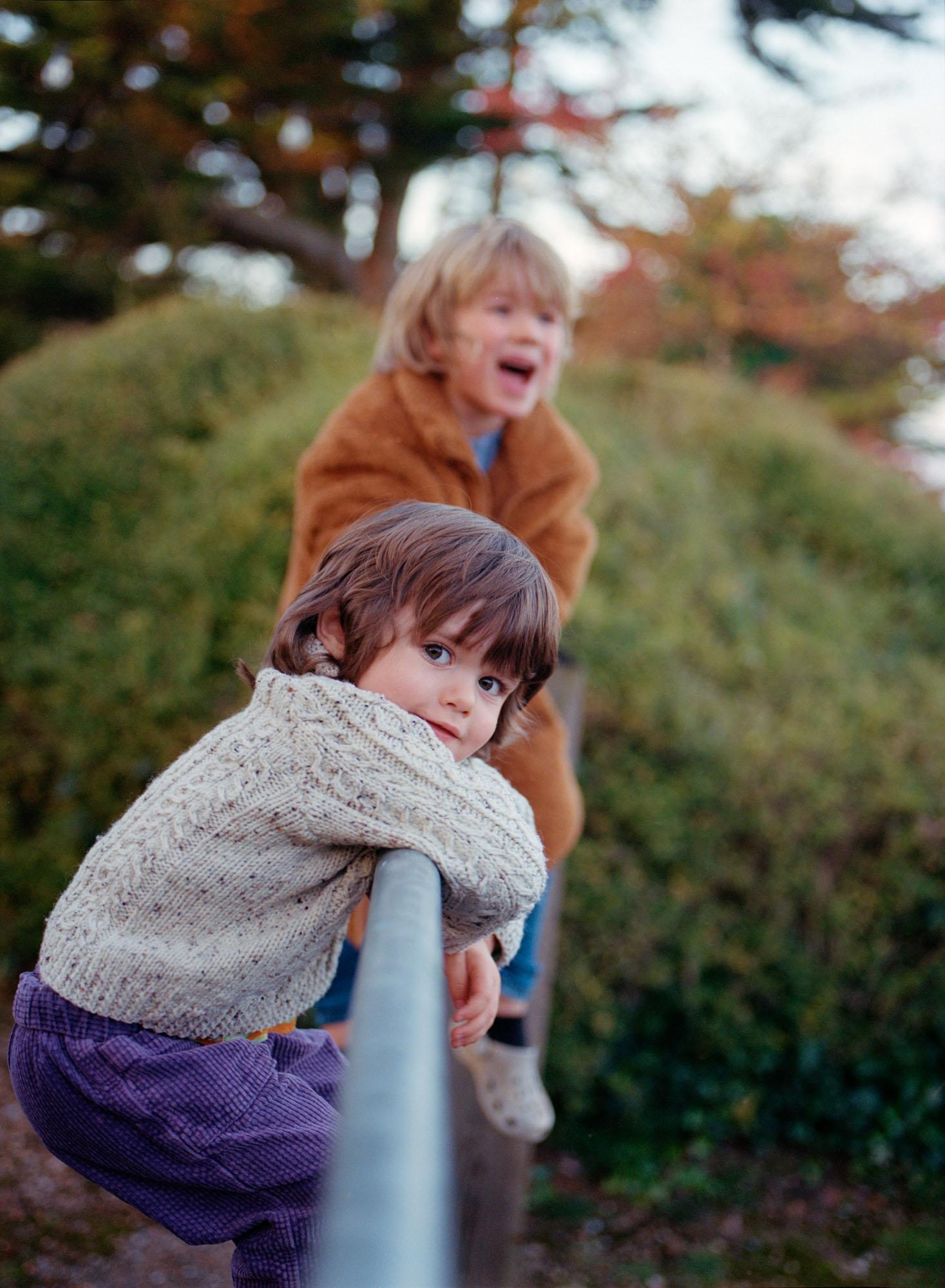 Two young children, likely siblings, are standing on a railing, looking out at something in the distance, both wearing casual, autumnal clothing.