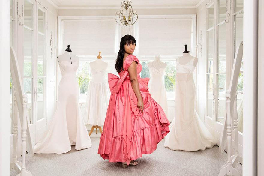 A woman in a pink ball gown stands in a room filled with white wedding dresses, surrounded by large windows and a chandelier.