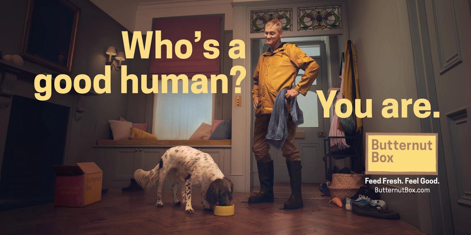 A man in a yellow jacket and boots standing in a dark room with a dog eating from a yellow bowl, with a butternut box in the background.