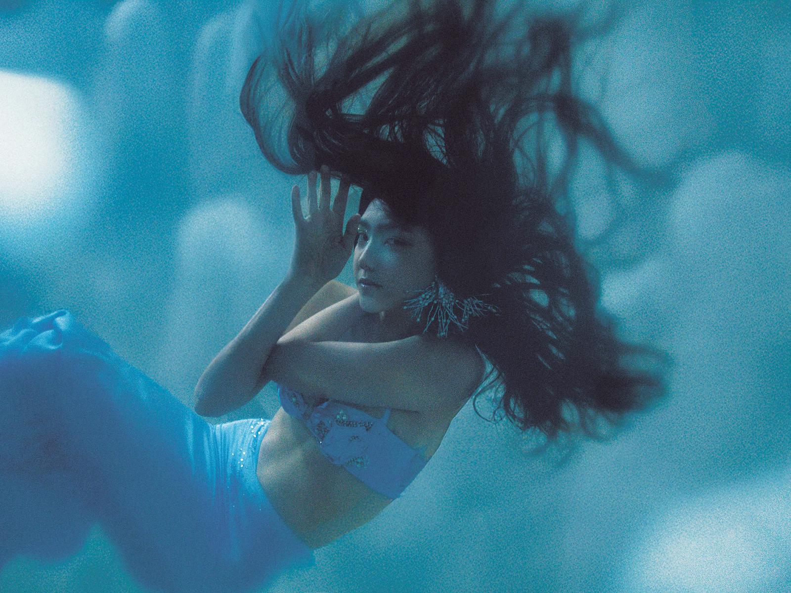 A woman in a mermaid tail swims underwater, surrounded by oceanic plants and fish.