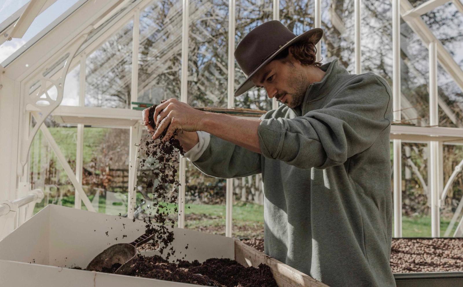 A man in a green sweater and a black hat is pouring soil into a large white container in a greenhouse.