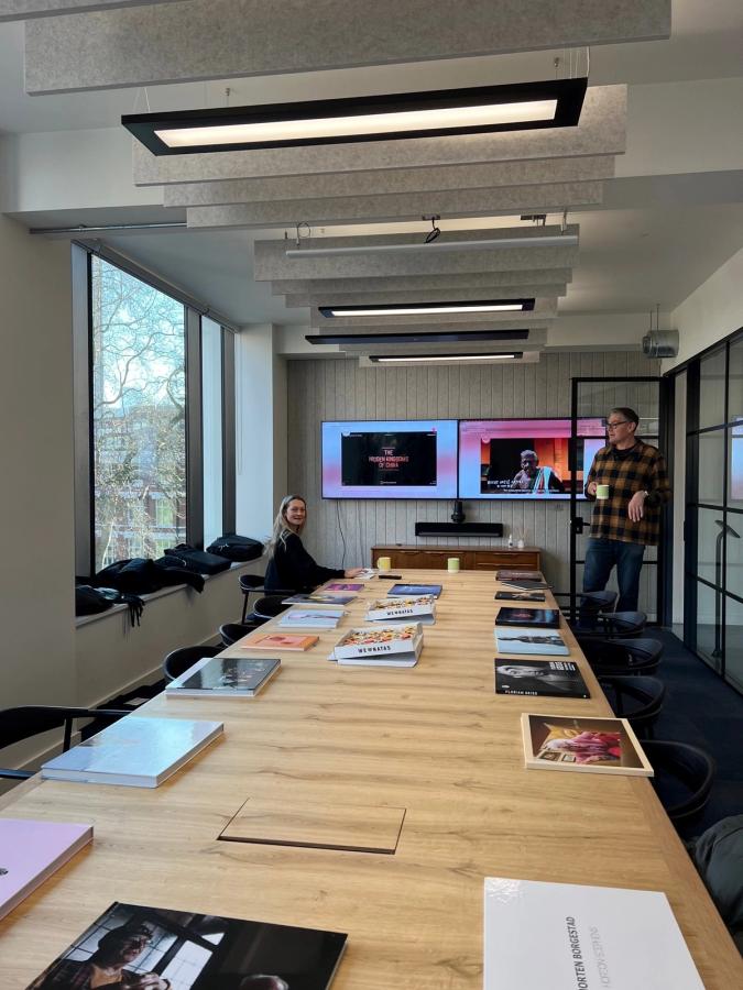 A conference room with a large table, a woman sitting at the head of the table, and a man standing at the end of the table, both with laptops and papers.