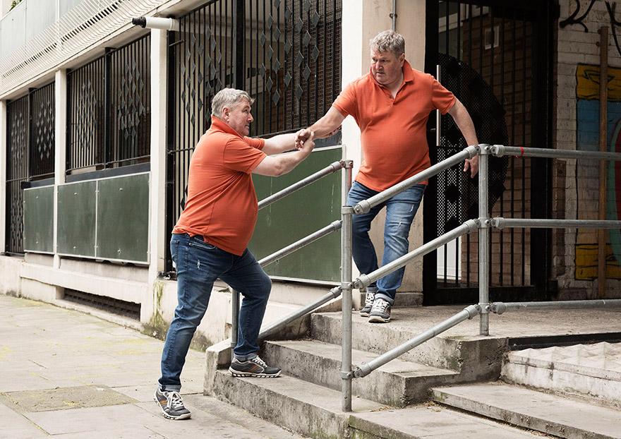 Two men with grey hair are descending concrete steps together, both wearing orange polo shirts and jeans.
