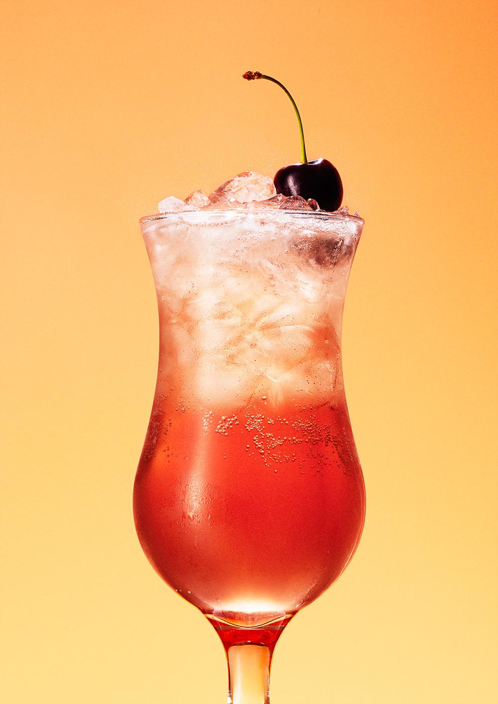 A glass of pink, fizzy cocktail with a cherry garnish, served in a tall, slender glass with a curved, fluted shape, and a long, thin stem.