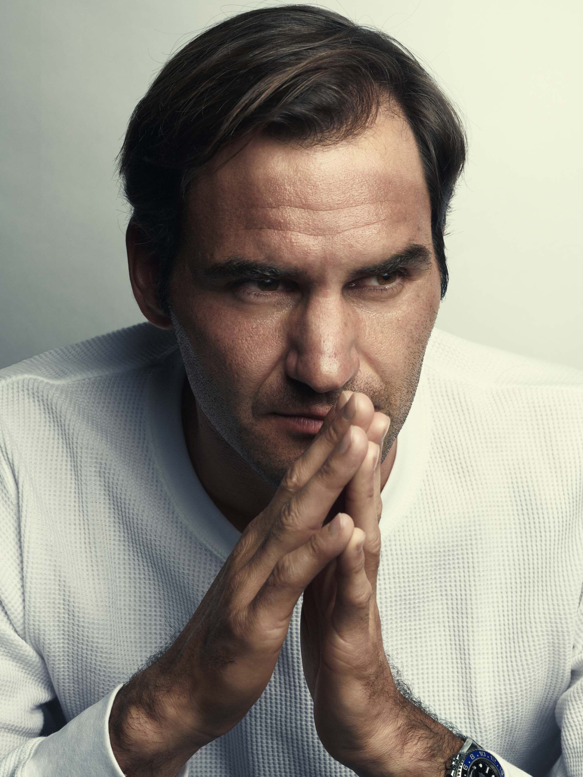 A man with dark hair and a white waffle-textured sweater, looking directly at the camera with his hands clasped together in front of his mouth.