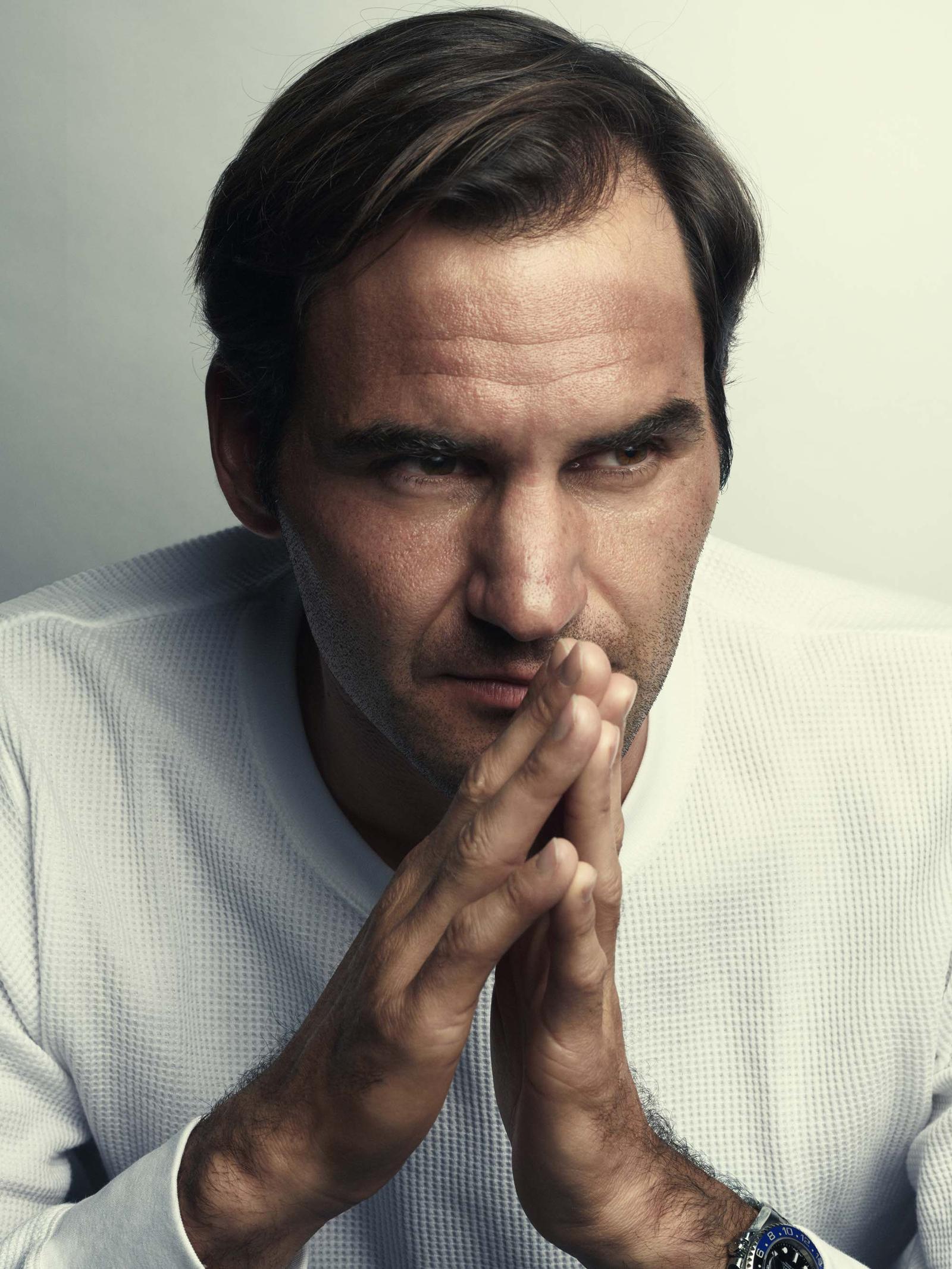 A man with dark hair and a white waffle-textured sweater, looking directly at the camera with his hands clasped together in front of his mouth.