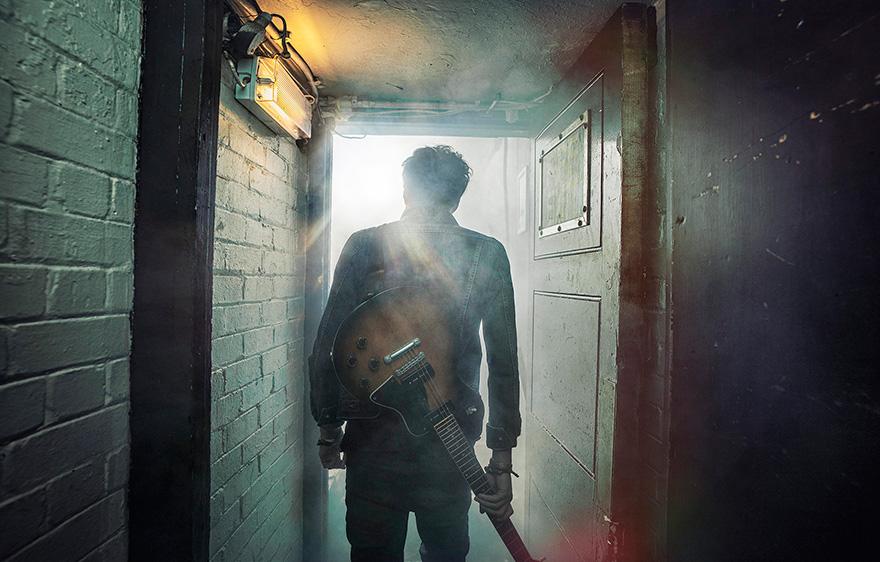 A man in a dark leather jacket and jeans, holding a guitar, standing in a dimly lit hallway with a door to his left and a wall of brick to his right.