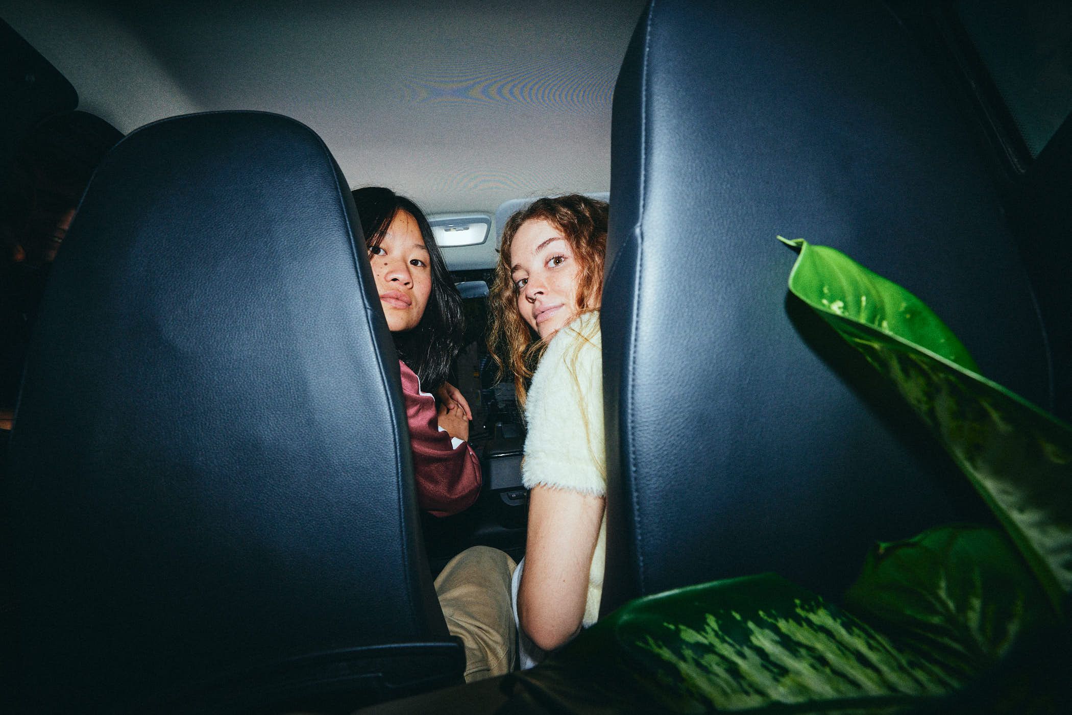 Two young girls, one with dark hair and the other with light hair, sit in the back of a car, looking back at the camera with a mix of curiosity and friendliness. The atmosphere.