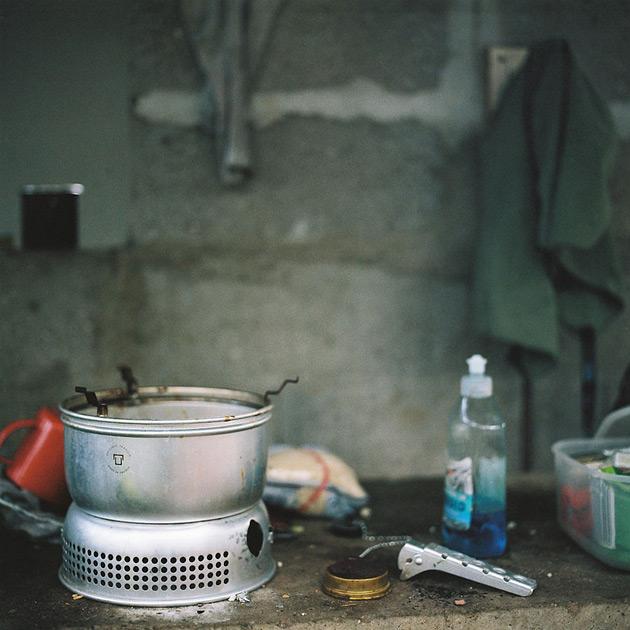 A small, portable, metal, camping-style, portable, gas-powered, cooking stove, with a metal, fuel container, on a concrete, outdoor, table, in a dimly lit, outdoor, environment.