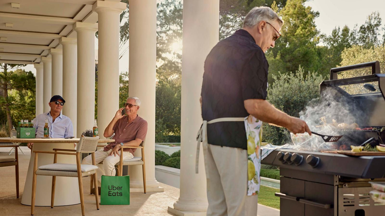 A man in a black shirt and white apron stands near a barbecue grill with two older men sitting at a nearby table.