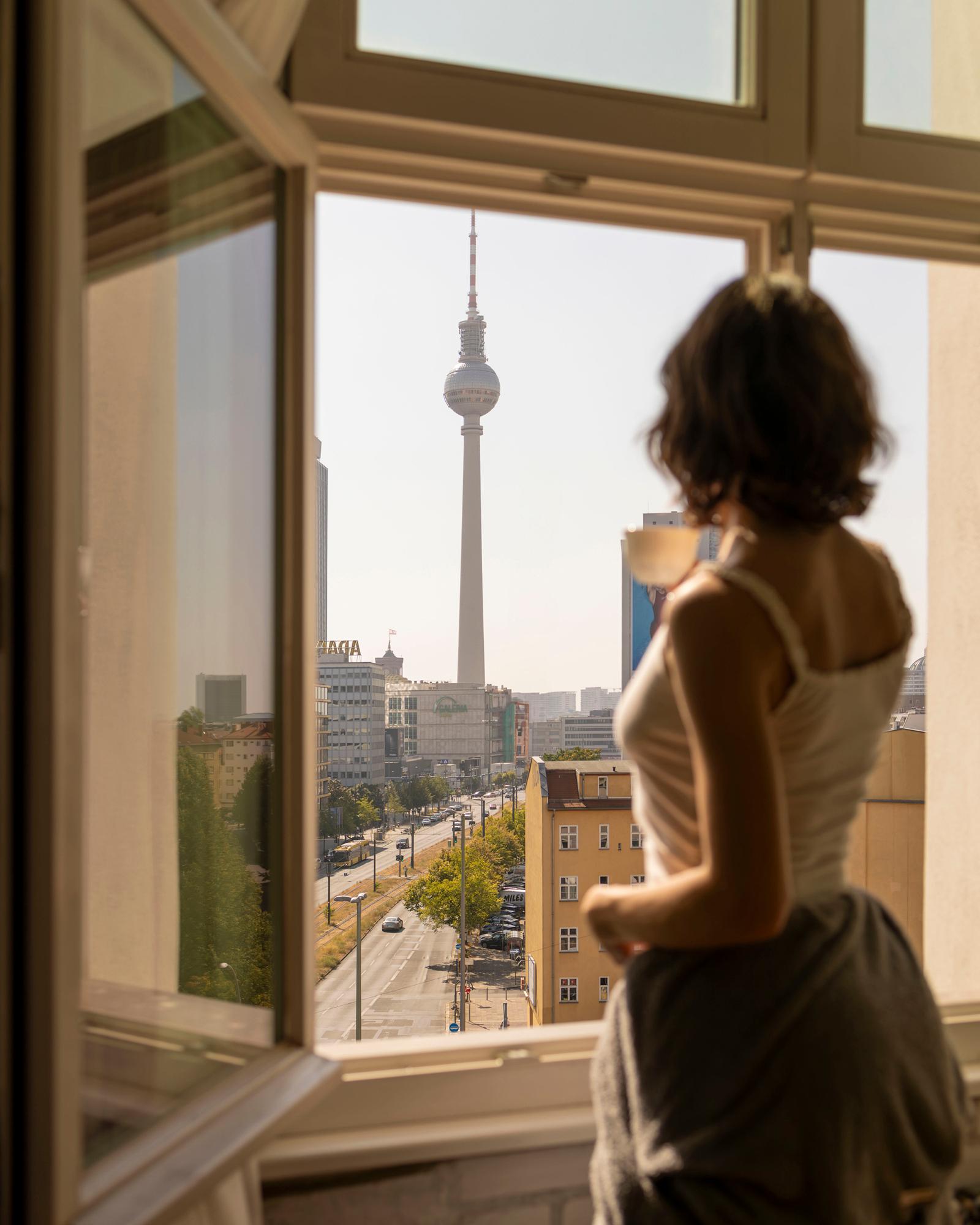 A woman in a white tank top and grey skirt is standing in front of a window, looking out at the Berlin skyline, with the Berlin TV Tower (Berliner ...