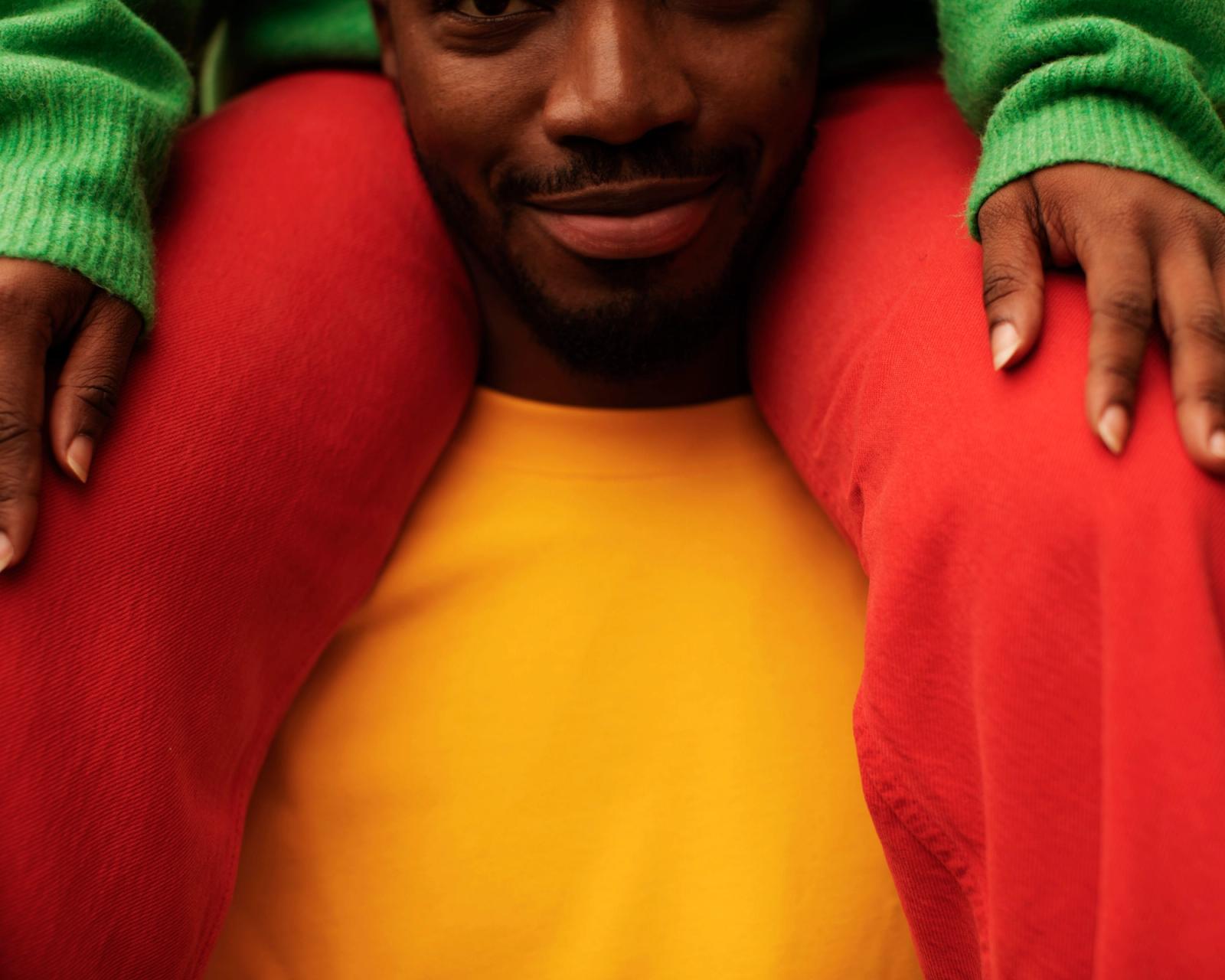 Two people sit on a couch, one in a yellow shirt and red pants with a green sweater, the other in a green sweater, both with hands on the couch.