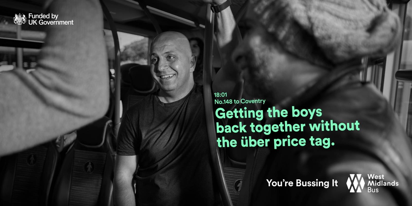 A man in a black t-shirt is smiling and waving at someone on a bus, with a blurred background of other passengers.